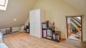 Um quarto com teto inclinado, um guarda-roupa branco, uma estante com livros e uma porta que dá para uma escada.