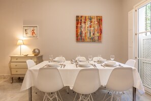 Un comedor con una mesa dispuesta para seis personas, sillas blancas, una lámpara, una mesita con un cuenco y un adorno, y un cuadro abstracto colorido en la pared.