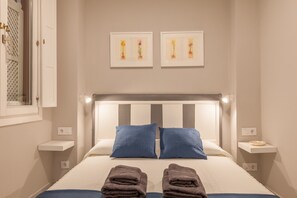 A hotel room with a bed, two pillows, a bedside table, a nightstand, a window, and two wall-mounted pictures. - City Centre Apartment with Balcony, Elevator and WiFi (Seville)
