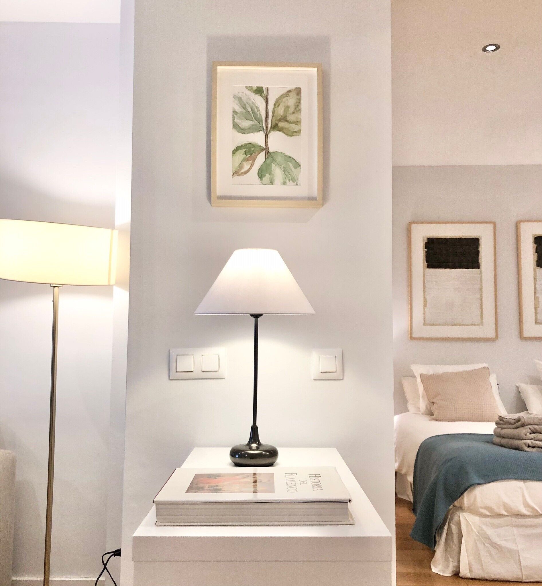 Un dormitorio con una cama, una mesita de noche con lámpara, un cuadro en la pared y un libro sobre la mesa.