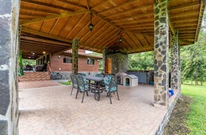 A covered patio area with a stone column structure, a table with chairs, and a view of a house in the background. - A country estate surrounded by nature with the best climate, just 2 minutes from the main road (El Valle de Antón)