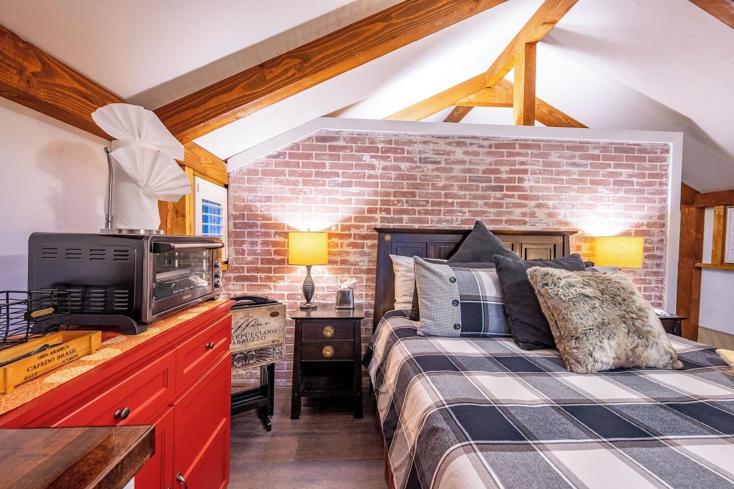 A bedroom with a brick wall, a bed with a plaid blanket, two bedside lamps, a red dresser, and a vintage radio.