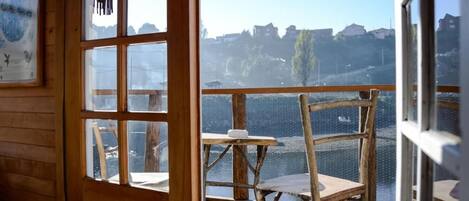 Una terraza de madera con una mesa y sillas, con vista a un cuerpo de agua y edificios a lo lejos.