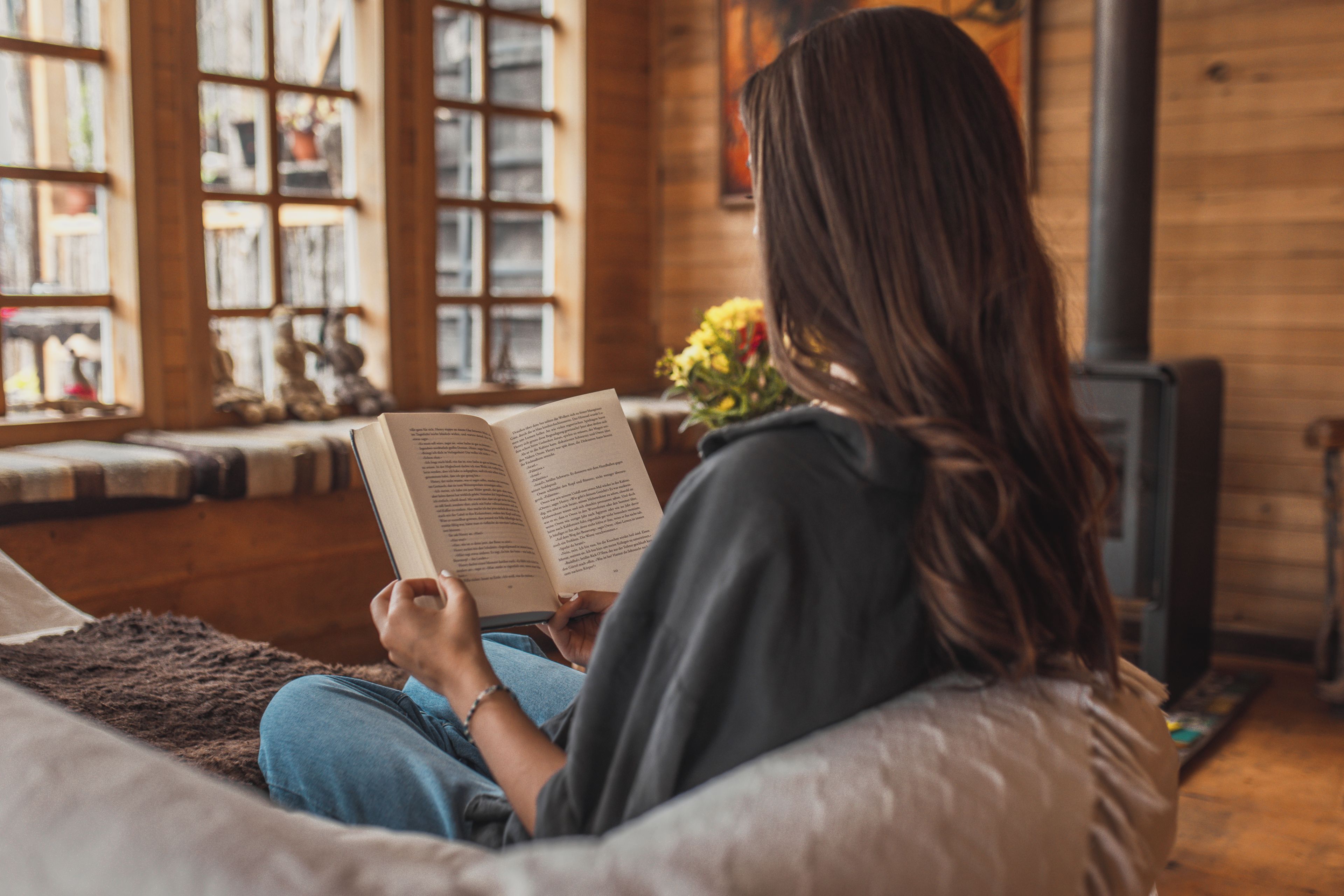 Une personne assise sur un canapé, lisant un livre dans une pièce où se trouvent une cheminée et une bibliothèque.