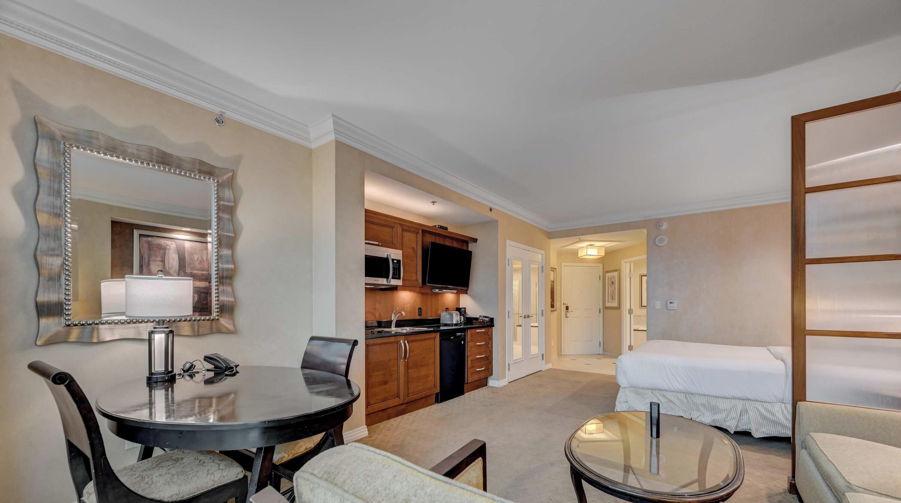 A hotel room with a dining table, a chair, a bed, a TV, and a kitchen area.