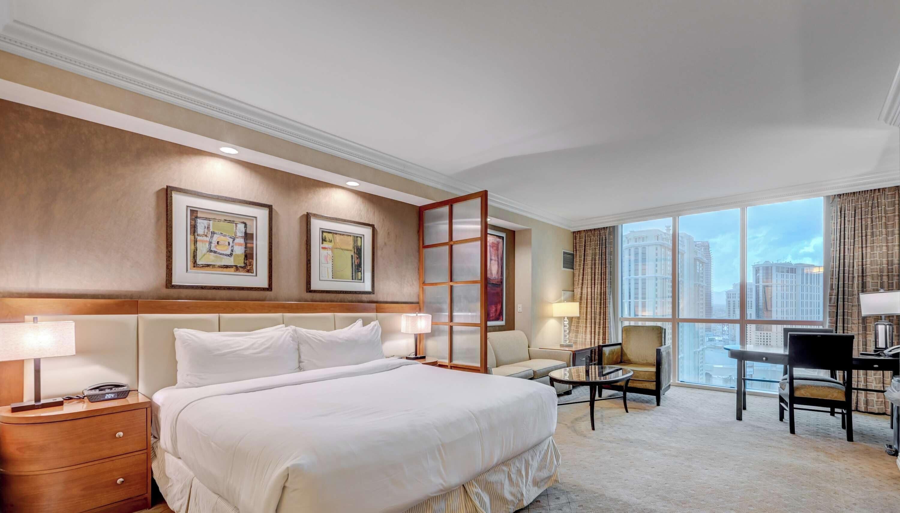 A hotel room with a large bed, a desk, a chair, and a view of the cityscape.