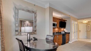 Quarto de hotel com mesa de jantar, um espelho grande, área de cozinha com armários de madeira, micro-ondas e TV de tela plana.