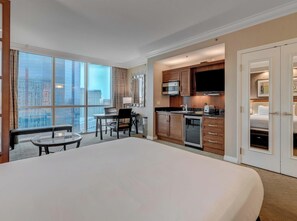 Een hotelkamer met een groot bed, een zithoek met een tafel en stoelen, een kitchenette met een magnetron en fornuis, en uitzicht op de stad door grote ramen.