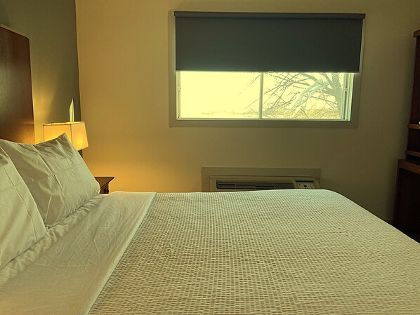 Una habitación de hotel con una cama, una mesita de noche, una lámpara, un ventanal con vistas y un equipo de aire acondicionado.