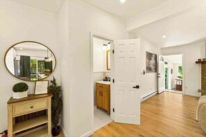 Une pièce spacieuse avec un plancher en bois, une porte blanche donnant sur une salle de bain, un miroir avec un cadre doré et une petite plante en pot posée sur une commode.
