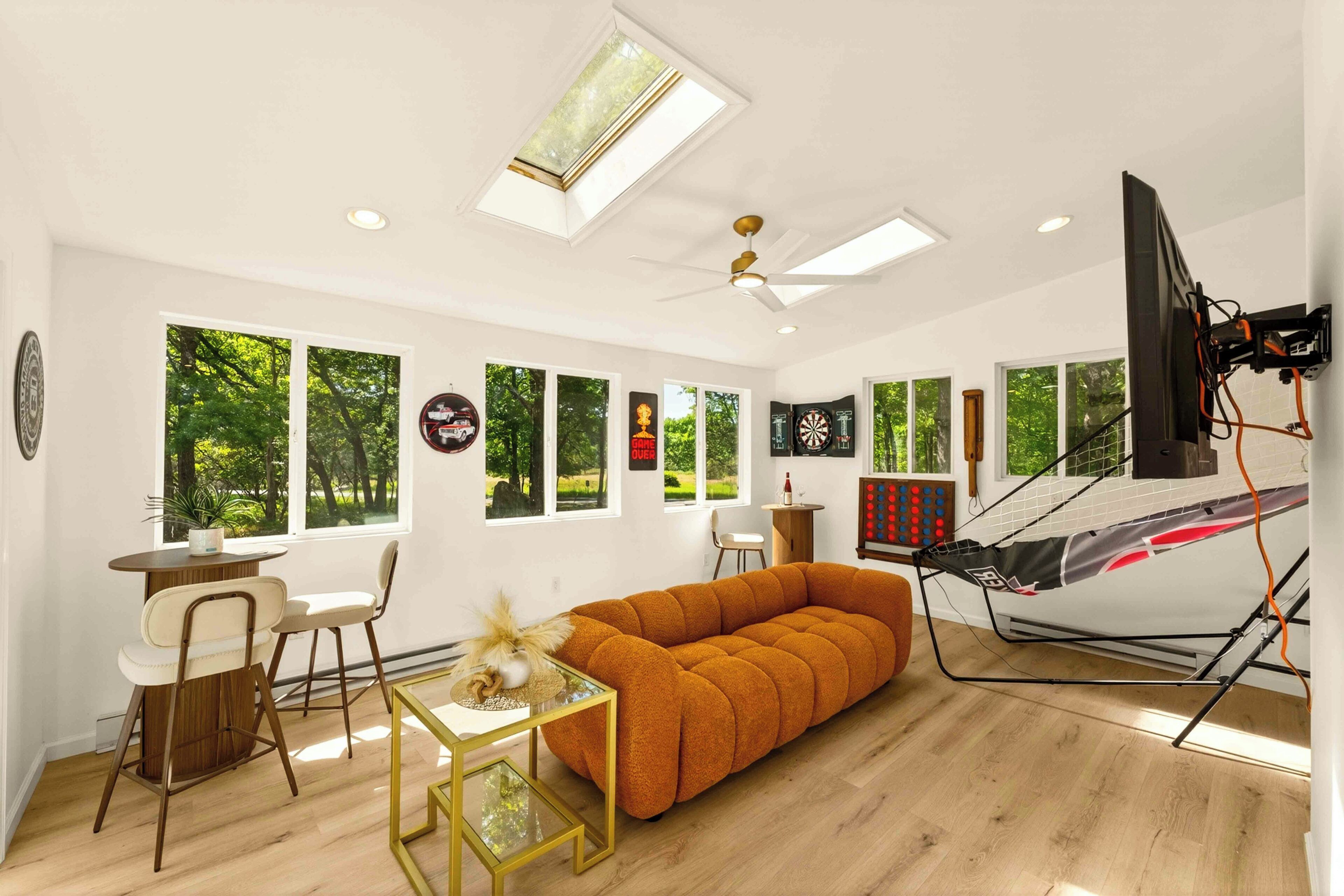 A modern living room with a sofa, a dining table, and chairs. There are large windows with a view of trees.