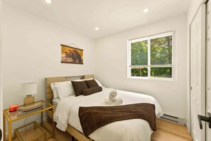 Une chambre à coucher comprenant un lit, une table de chevet, une lampe, une fenêtre avec des stores et un tableau au mur.
