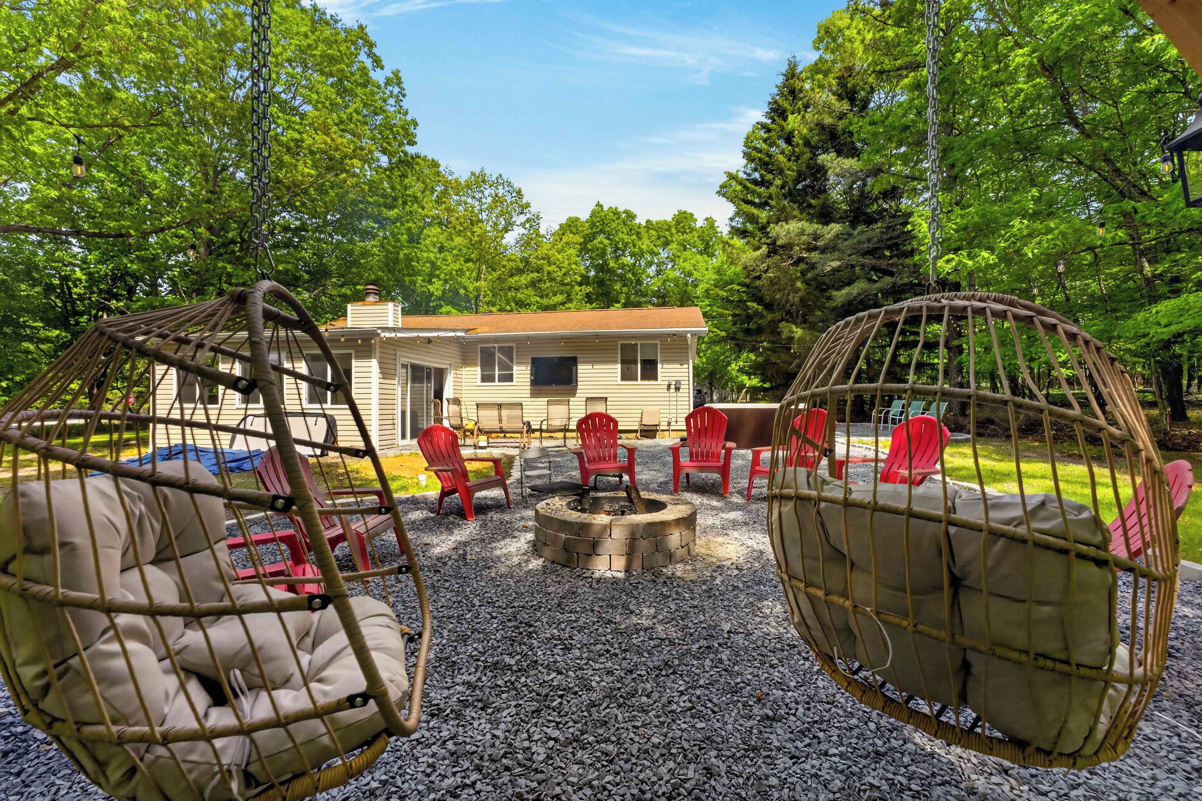 A rustic outdoor seating area with wicker chairs and a fire pit, surrounded by gravel and trees.