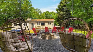 A rustic outdoor seating area with wicker chairs and a fire pit, surrounded by gravel and trees.
