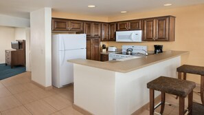 Una cocina con armarios de madera, un refrigerador, un microondas, una estufa y un lavamanos. Hay dos taburetes y una mesita.