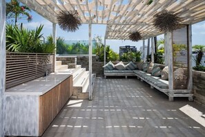 Un espace extérieur abrité, aménagé avec un banc en bois, un canapé rembourré et une pergola décorée de guirlandes.