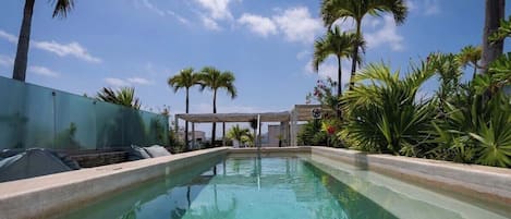 A rectangular swimming pool with clear water, surrounded by palm trees and a pergola structure.
