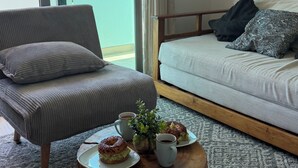 Ein modernes Wohnzimmer mit einer Couch, einem Couchtisch mit Gebäck und Blick auf Palmen durch große Fenster.