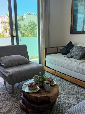 A modern living room with a sofa, a coffee table with pastries, and a view of palm trees through large windows. - LUXURY Apartment. 1 block from 5th Avenue (Playa del Carmen)