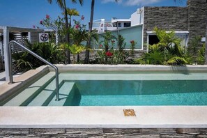 A swimming pool with a metal staircase leading down to the water, surrounded by tropical plants and a building with a stone facade. - LUXURY Apartment. 1 block from 5th Avenue (Playa del Carmen)