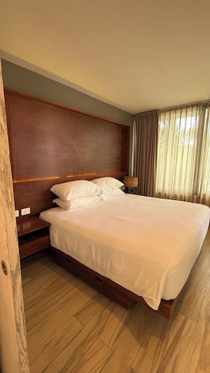 A hotel room with a large bed, wooden headboard, a nightstand, a lamp, and a window with curtains. - LUXURY Apartment. 1 block from 5th Avenue (Playa del Carmen)