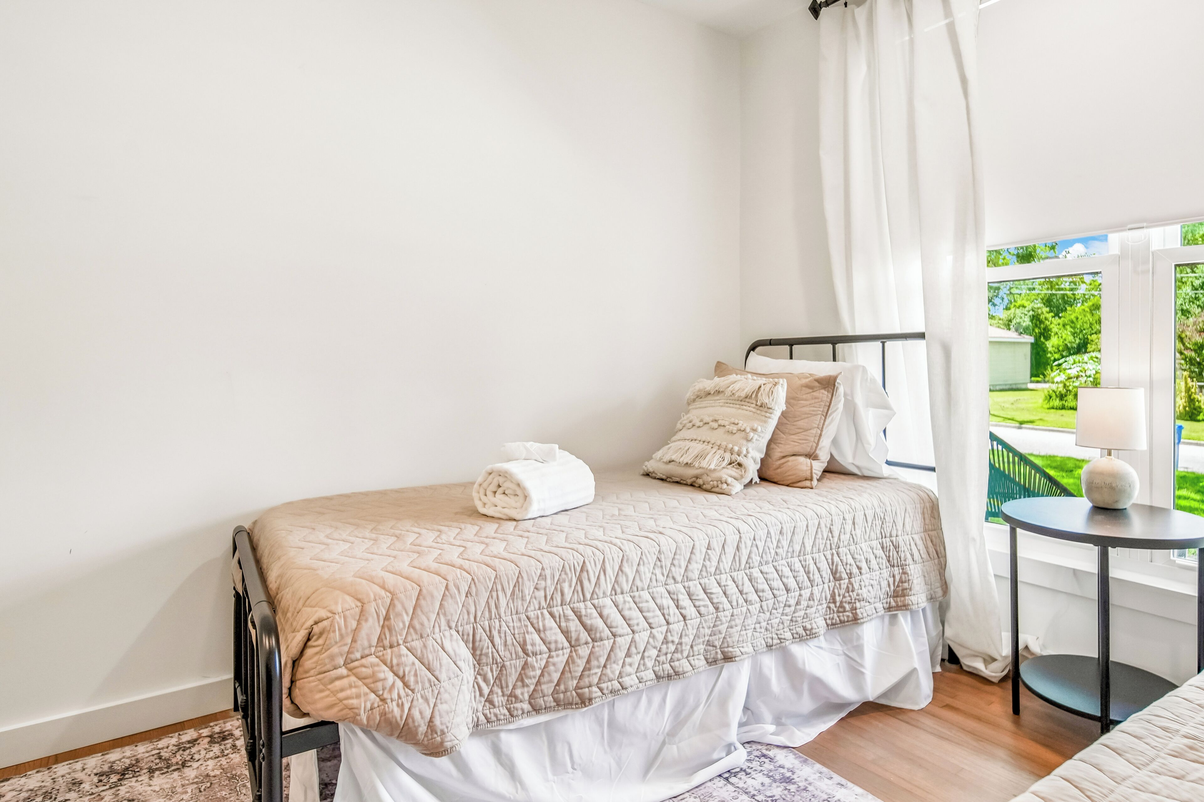 A bedroom with a bed, pillows, a rolled towel, a small round table with a lamp, and a view of greenery outside.