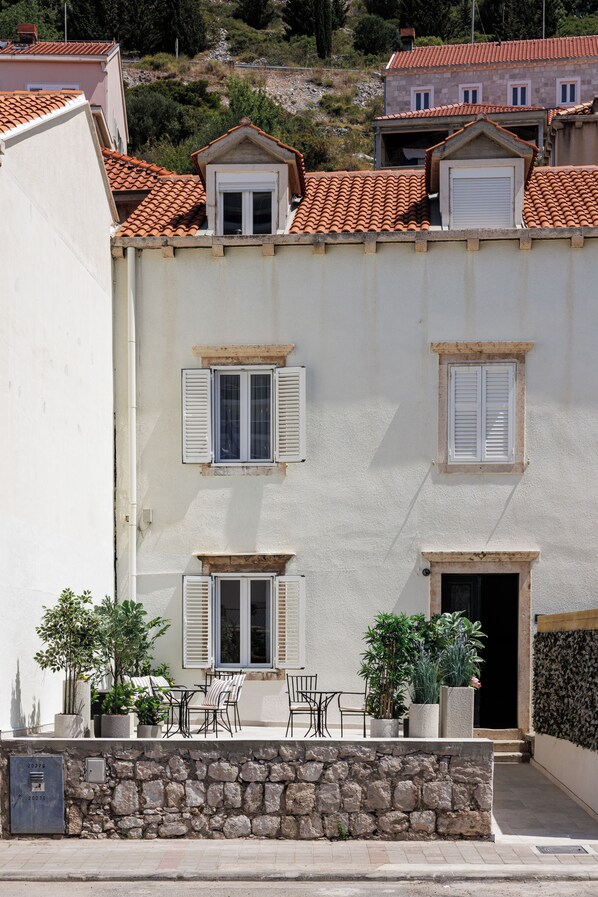 A building with a stone facade, multiple windows with shutters, and a small outdoor seating area with chairs and potted plants.