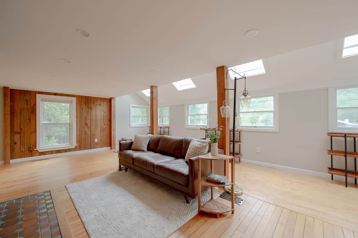 Een ruime kamer met een groot raam, een houten vloer, een bruine leren bank en een klein bijzettafeltje met een lamp.