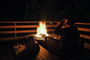 Une personne est assise à une table en bois, devant elle se trouvent un verre, un bol, et on aperçoit un foyer au loin.