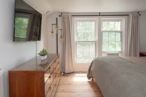 Une chambre à coucher avec un lit, une commode, un téléviseur fixé au mur et une fenêtre avec des rideaux.