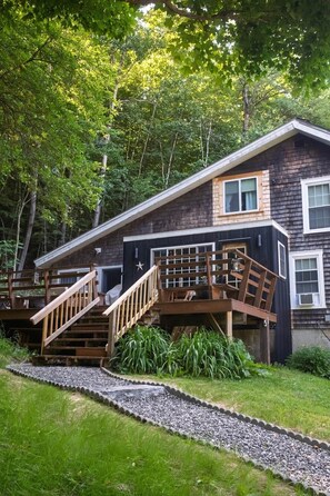 Une cabane à deux étages dotée d’une terrasse en bois, d’un escalier et d’un chemin de gravier menant à l’entrée.