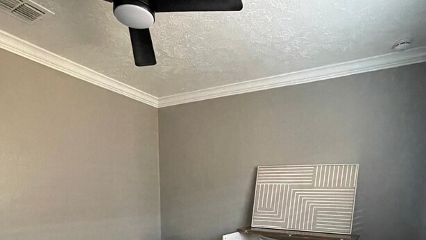 Una camera da letto con un letto singolo, una sedia, una ventola a soffitto e un'opera d'arte appesa al muro.