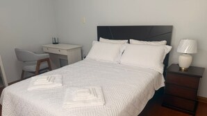 Uma cama bem arrumada com cabeceira, uma mesa de cabeceira com abajur e uma cadeira em um quarto com paredes em tons claros.
