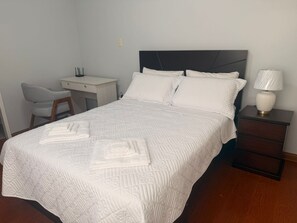 Ein ordentlich bezogenes Bett mit Kopfteil, ein Nachttisch mit Lampe und ein Stuhl in einem Zimmer mit hellen Wänden.