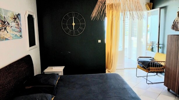 Ein Schlafzimmer mit Bett, einer Wanduhr, einem Fenster mit Vorhängen und Blick ins Freie.