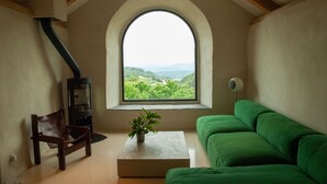 Ein Zimmer mit einem grünen Sofa, einem Holzstuhl, einem kleinen Tisch mit einer Pflanze, einem Kamin und einem Fenster mit Blick auf Grünflächen und Hügel.