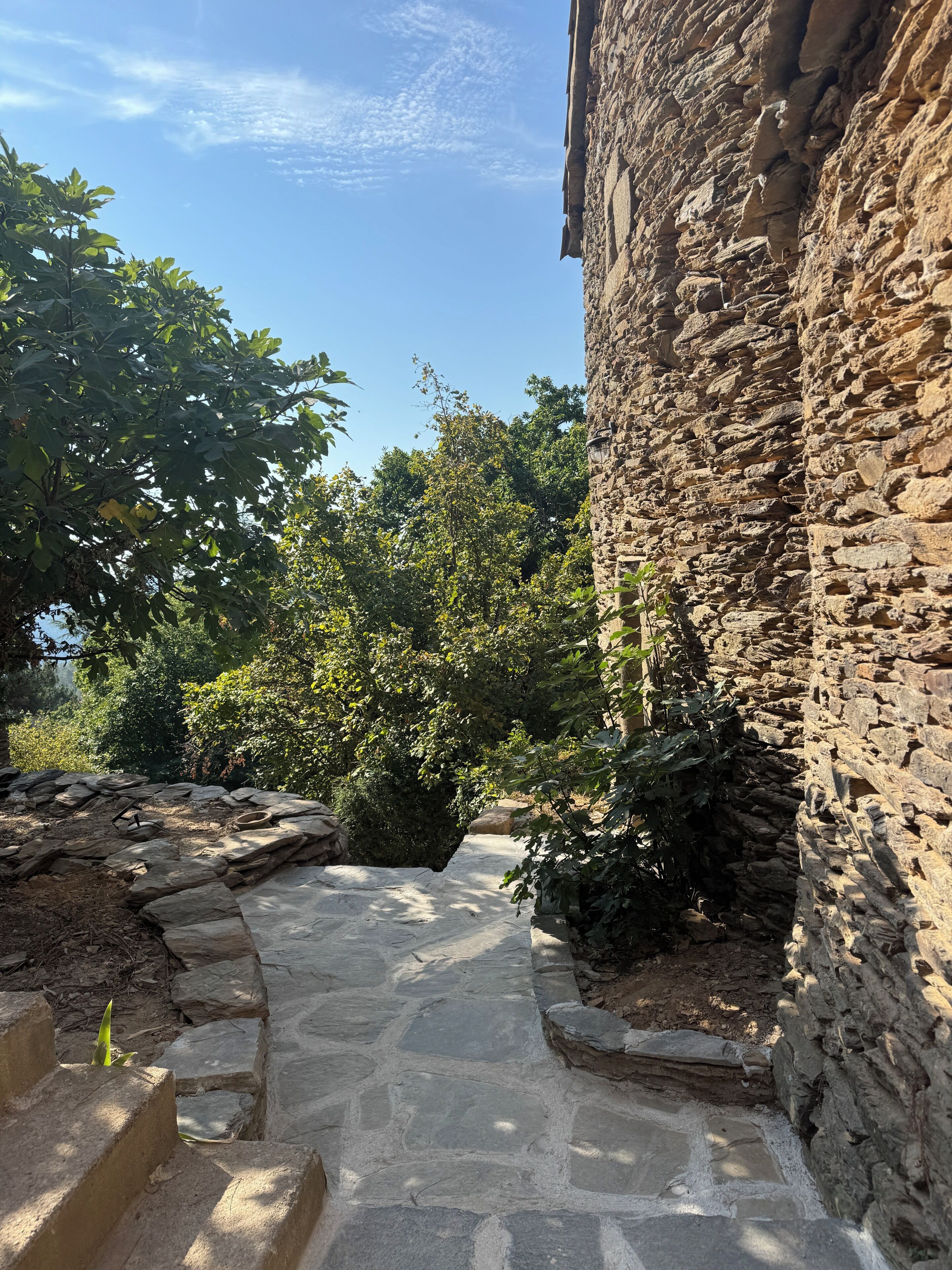 A stone pathway leading to a rustic building with a stone wall and greenery around.