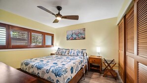 A bedroom with a bed, a bedside table, a nightstand, a ceiling fan, a window with blinds, and a wooden wardrobe.