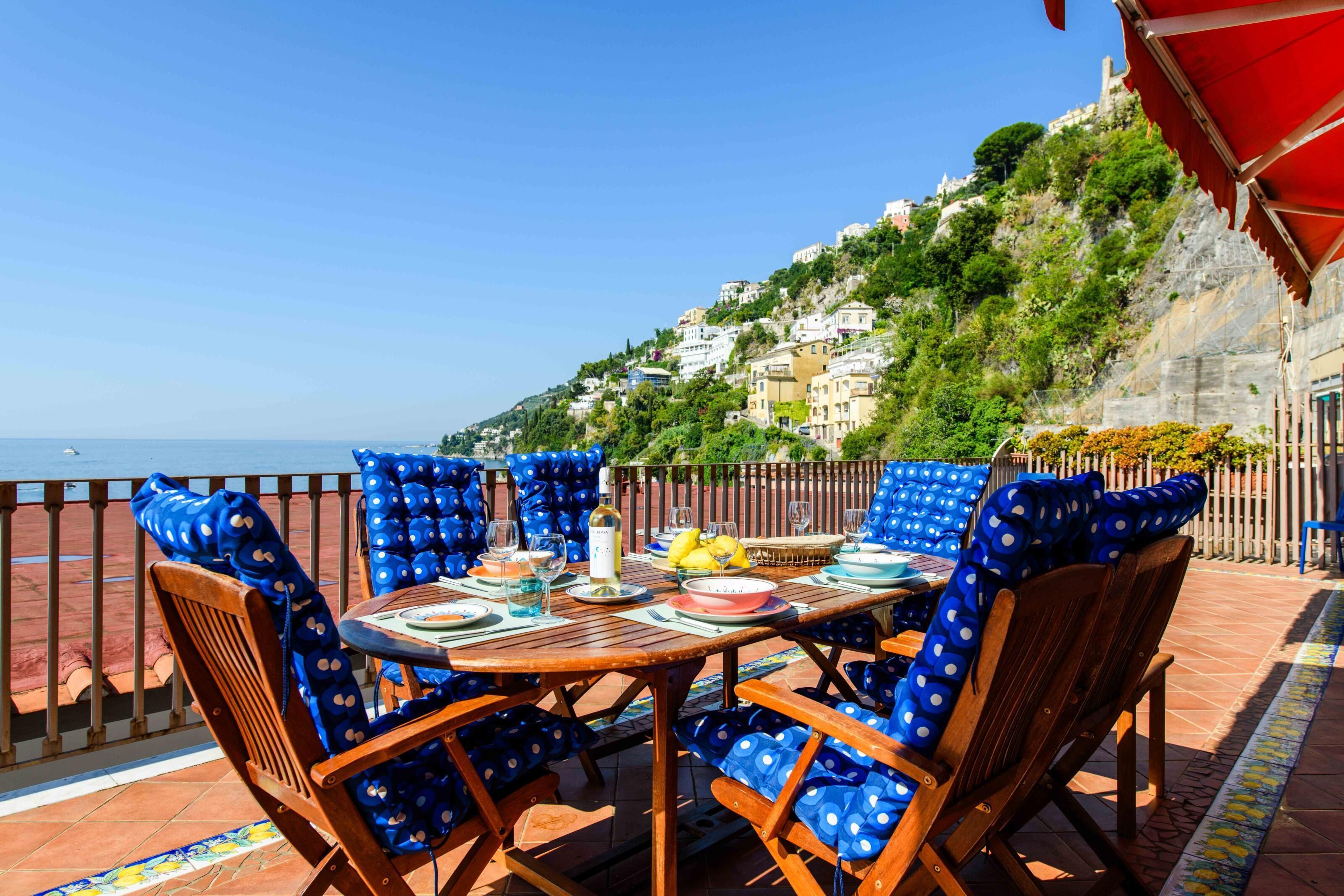 Mesa lista para comer con sillas, vista al mar y edificios en una colina.