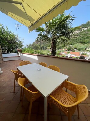 Eine Terrasse im Freien mit Tisch und Stühlen, einem gelben Sonnenschirm und Blick auf eine Hügelslandschaft mit Vegetation und Gebäuden.