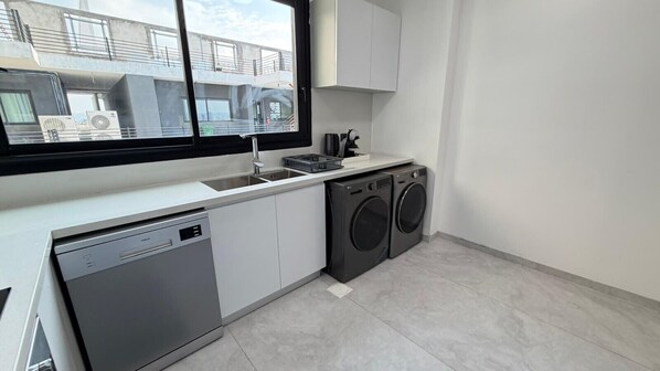 A compact kitchen with a dishwasher, two washing machines, a sink, and a window overlooking a building.