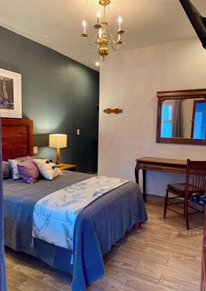 A bedroom with a bed, bedside table, lamp, wooden chair, and a mirror on the wall.