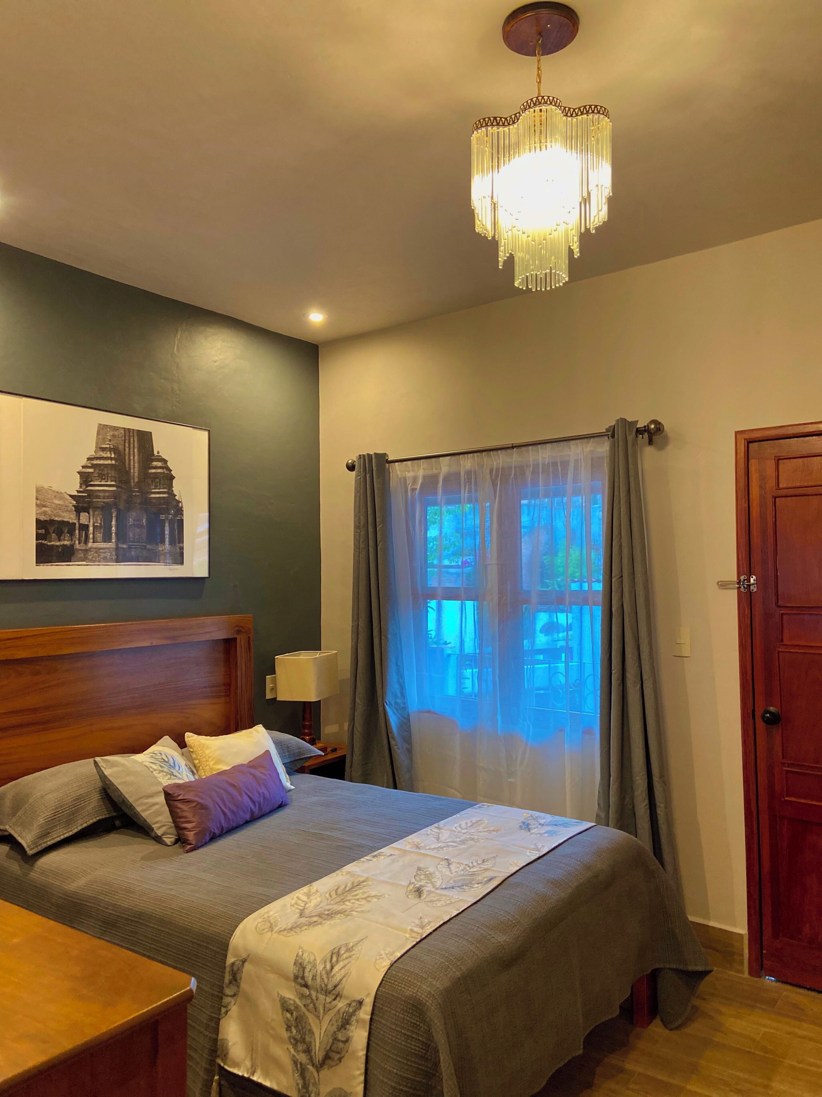 A bedroom with a bed, a nightstand, a lamp, a window with curtains, a door, and a chandelier.