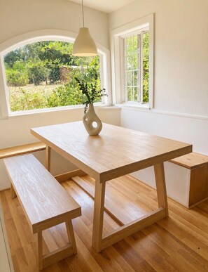 A minimalist dining area with a wooden table and benches, a hanging light fixture, and a view of greenery through a window. - Off-Grid Hidden Oaks Private Retreat (Atascadero)