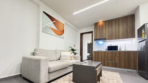 A modern living room with a sofa, coffee table, and a rug. A kitchen with wooden cabinets and appliances is visible in the background.