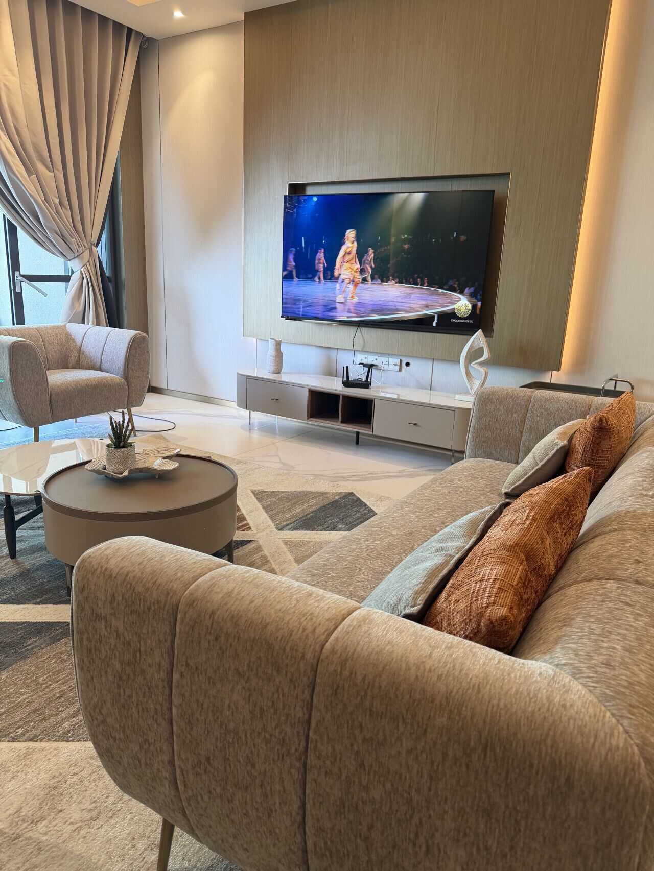 A modern living room with a flat-screen TV mounted on the wall, a beige sofa, a coffee table, and a patterned rug.