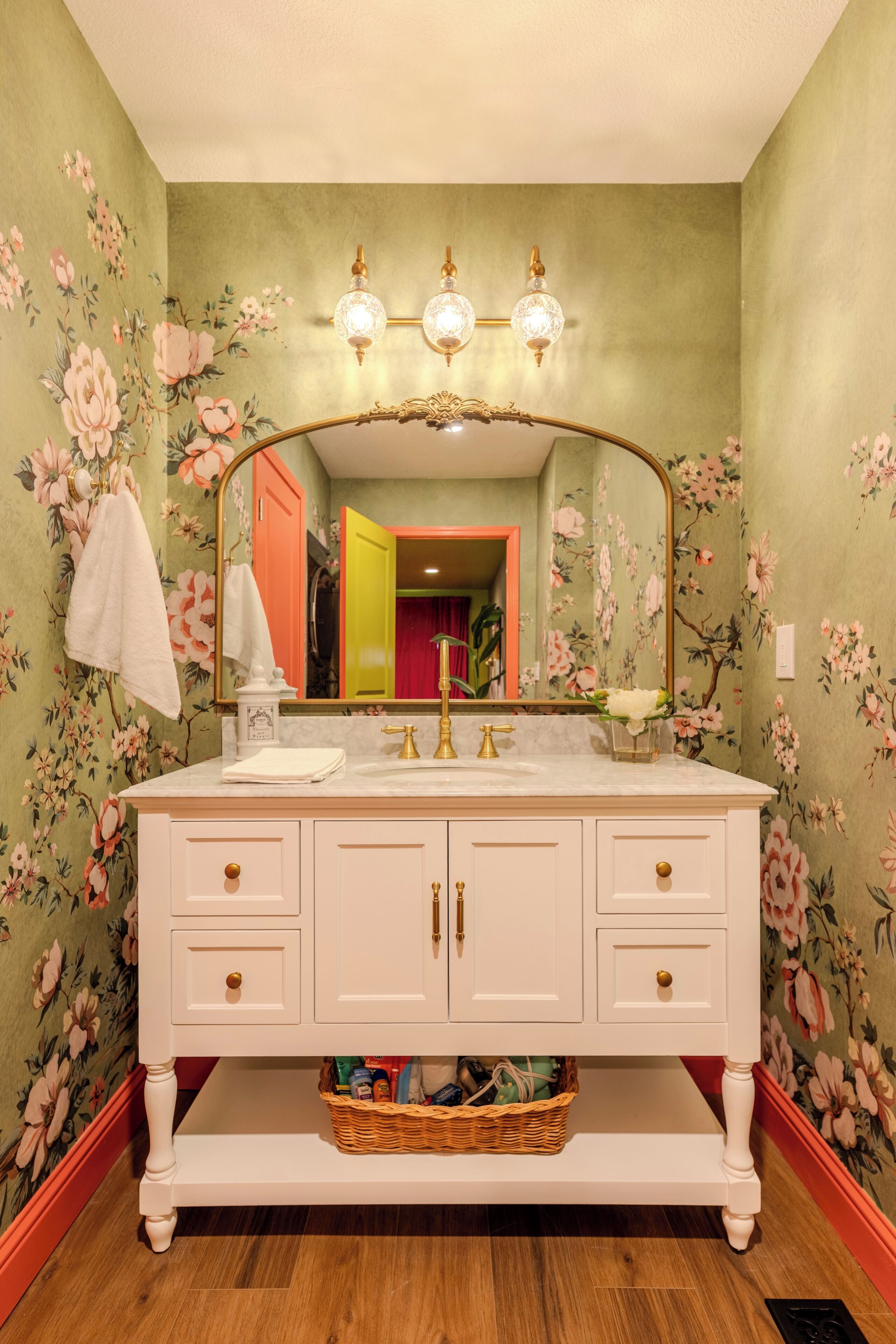 A bathroom with a floral wallpaper, a white vanity with gold fixtures, and a large mirror above the sink.