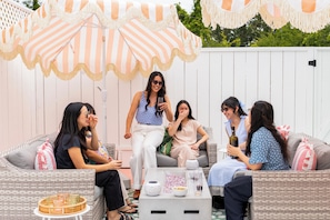 A group of people seated on outdoor wicker furniture, engaged in conversation, with a table in front of them holding drinks and a speaker. - The Gilded Bungalow • Walk to Town Green • Hot Tub • Firepit • Pink Kitchen (Windsor)