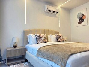 A modern hotel room with a bed, bedside table, lamp, air conditioning unit, and a wall-mounted artwork.