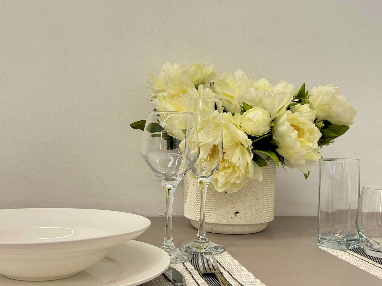 Een gedekte tafel met een wit bord, twee wijnglazen, een vaas met witte bloemen en een helder glas.